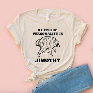 An off-white graphic t-shirt with a black printed text and raccoon illustration, styled with a knot at the bottom and paired with light blue jeans on a solid pink background.